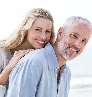 Half-body photo of a blonde middle-aged woman leaning on the back of a gray-haired man with a beard. Both of them are leaned forward and looking to their right and smiling - for information on teeth whitening from your dentist. There is pale-blue water in the lower background.